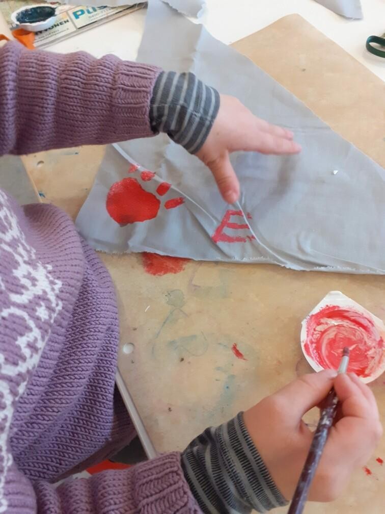 Lapsi maalaamassa punaisella kangasmaalilla harmaaseen huiviin.